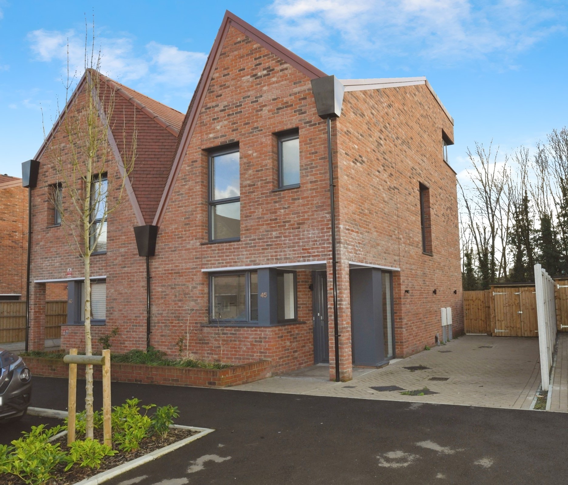 A semi-detached property with red brickwork and a quirky slanted roof. The two-storey house has driveway parking, and features dark window and door frames