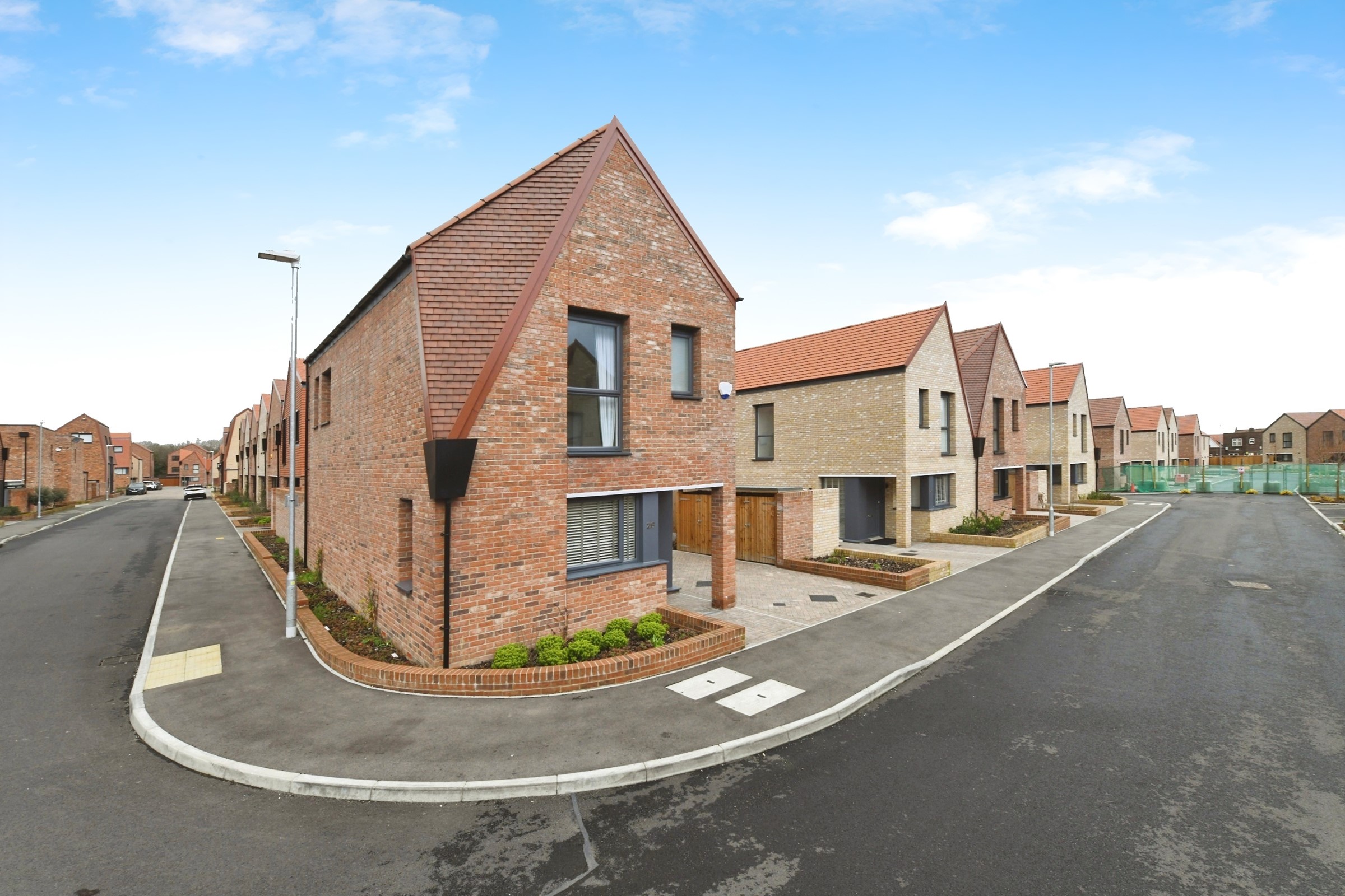 A detached, corner property with red brickwork and a red roof with dark window and door frames. The property features a carport and driveway parking and has a quirky, slanted roof