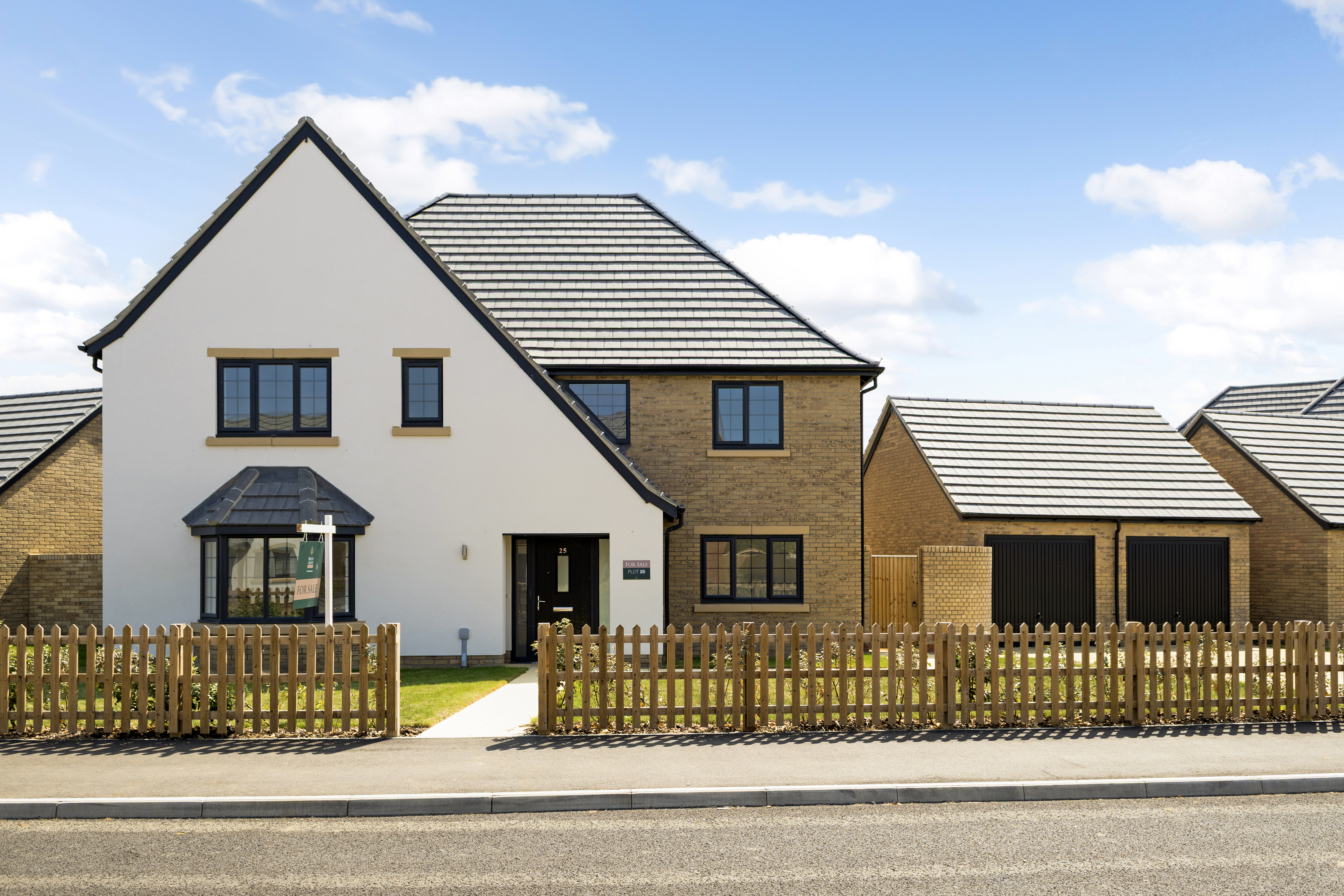  Contemporary two-story home with white and brick façade, large windows, and a dark grey tiled roof. Features include a manicured front lawn, wooden picket fence, and a visible "For Sale" sign. A detached double garage sits beside the house
