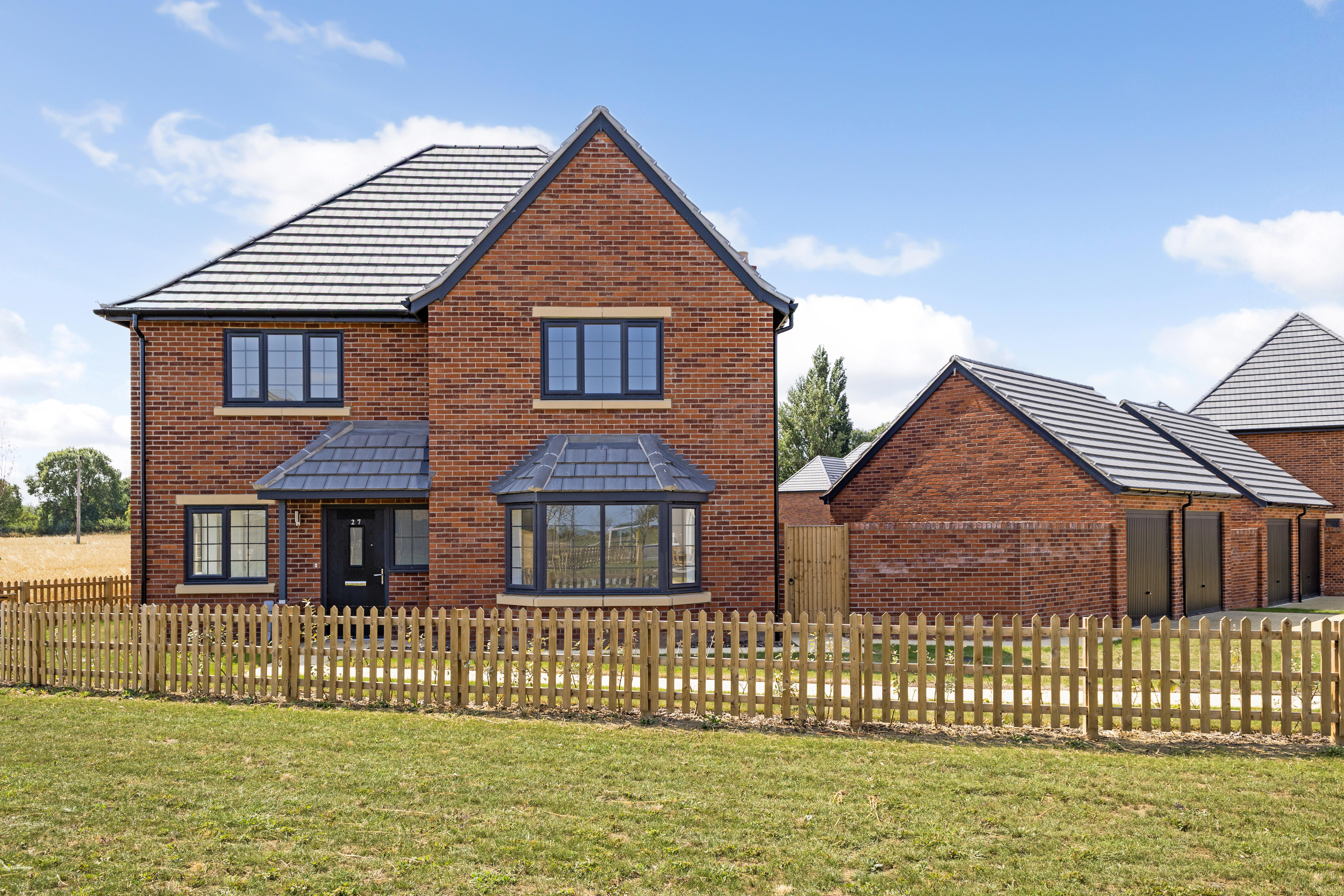 Elegant two-story brick home with grey tiled roof, bay window, and spacious front-facing windows. Includes a detached garage, a neat front garden bordered by a wooden picket fence