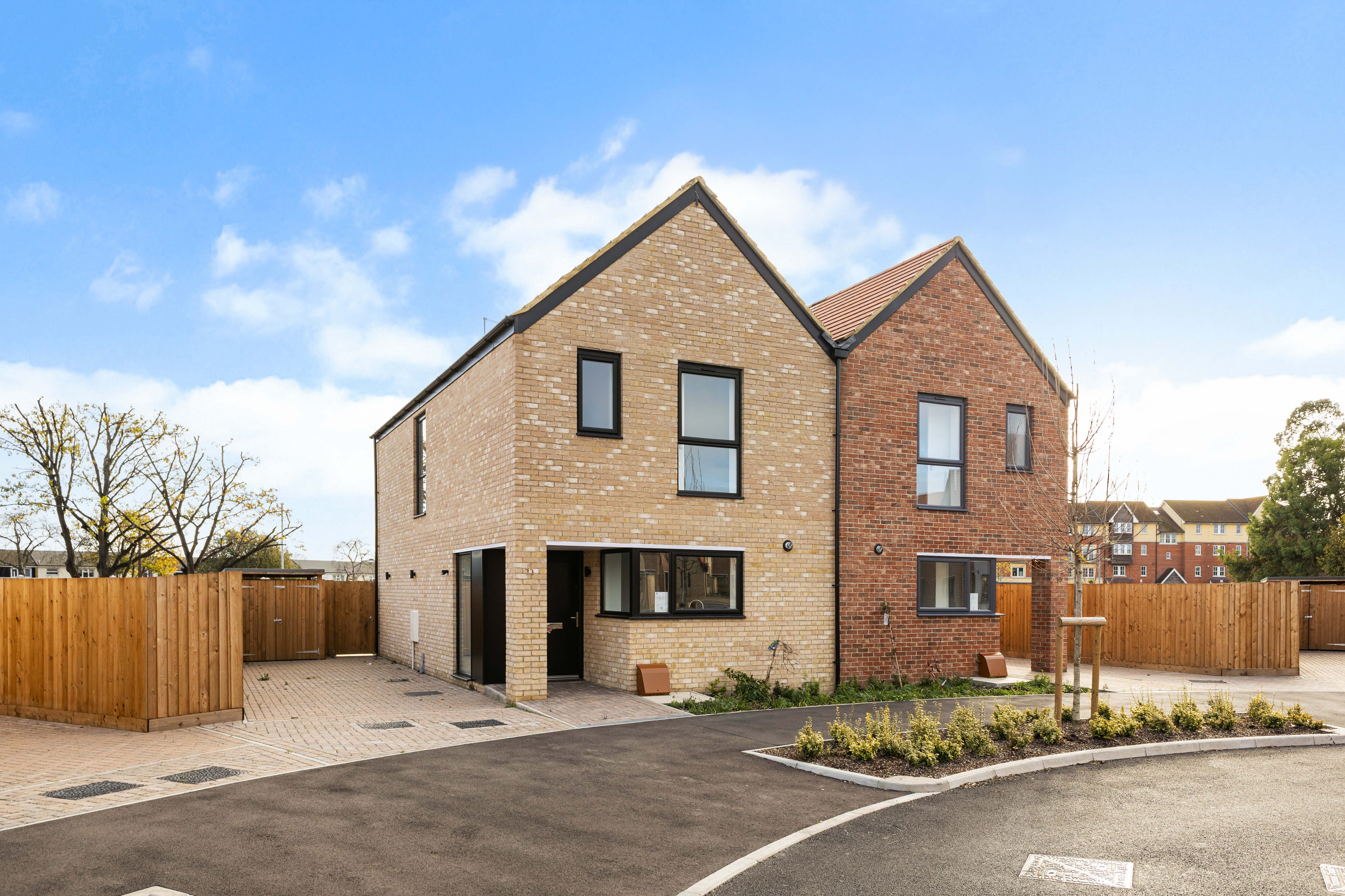 Modern two-storey semi-detached house with light and red brick exterior, black-framed windows, and a paved driveway, set in a residential development under a clear blue sky