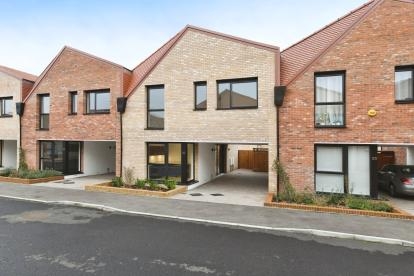Front of a mid-terrace property with light-coloured brickwork, dark window and door frames and a red roof. Carport parking and neat plant beds are at the front of the property.