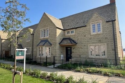 A stone two-story house with a pitched roof and multiple windows, a 'For Sale' sign on the lawn, and a tidy garden under a clear blue sky.