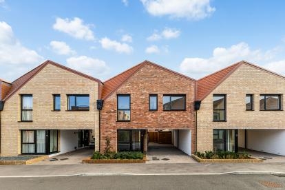 Row of modern two-storey terraced houses with light and red brick exteriors, pitched roofs, black-framed windows, and covered driveways in a new residential development under a partly cloudy sky.