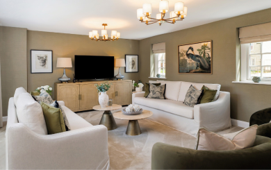 A stylish living room with two white sofas and two green armchairs arranged around a circular coffee table. The room has beige walls, three windows with Roman shades, and a large flat-screen TV mounted above a wooden cabinet. Two framed artworks and table lamps sit on either side of the TV. Two elegant chandeliers hang from the ceiling, enhancing the room’s sophisticated feel.