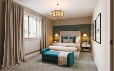 A modern, softly lit bedroom with a double bed centred against a green panelled feature wall. The bed has neutral bedding with teal cushions and a matching teal bench at the foot. Two bedside tables with warm pendant lights sit on either side. Large curtains frame a window on the left, and framed artwork hangs on the right wall.