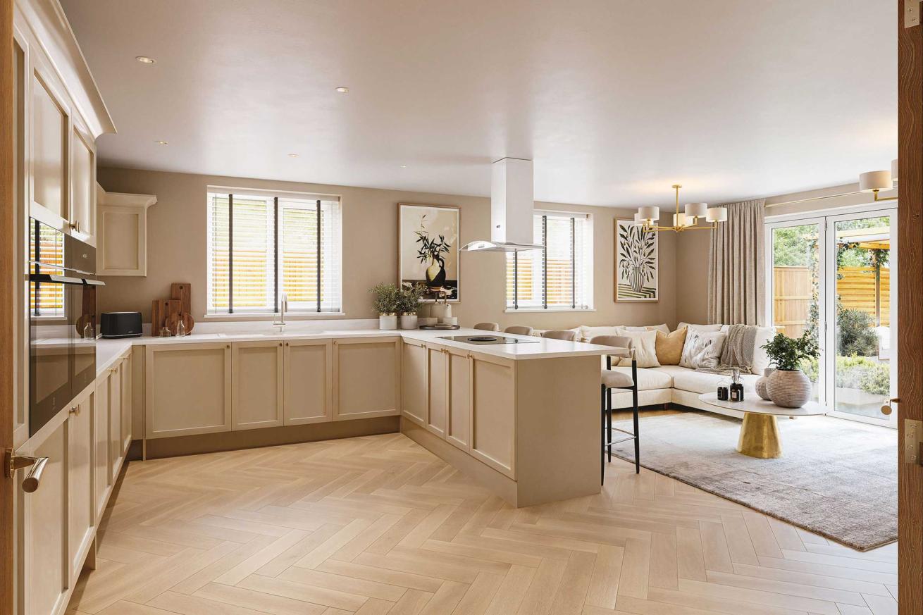 Spacious open-plan kitchen with cream cabinetry, a central island with integrated hob and bar stools, light wood herringbone flooring, and a cozy seating area with a sofa and coffee table near glass doors leading to a garden.