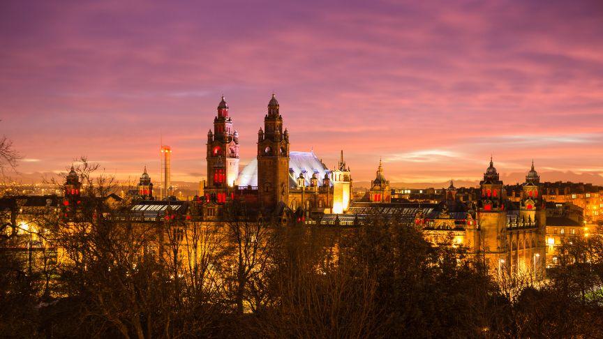 Panoramic view of Kelvingrove Art Gallery and Museum in Glasgow at sunset, with a colourful sky of purple, pink, and orange. The building is warmly lit, showcasing its towers and domes, with trees in the foreground