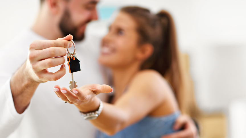 A man and woman embracing one another, gazing lovingly into each others' eyes. In the foreground the man is handing the woman a house key with a house shaped keyring attached.