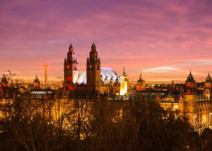 Panoramic view of Kelvingrove Art Gallery and Museum in Glasgow at sunset, with a colourful sky of purple, pink, and orange. The building is warmly lit, showcasing its towers and domes, with trees in the foreground