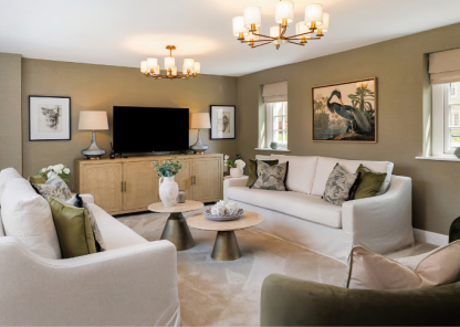 A stylish living room with two white sofas and two green armchairs arranged around a circular coffee table. The room has beige walls, three windows with Roman shades, and a large flat-screen TV mounted above a wooden cabinet. Two framed artworks and table lamps sit on either side of the TV. Two elegant chandeliers hang from the ceiling, enhancing the room’s sophisticated feel.