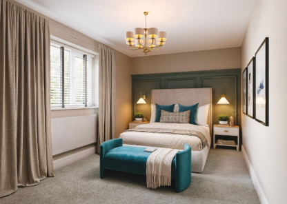 A modern, softly lit bedroom with a double bed centred against a green panelled feature wall. The bed has neutral bedding with teal cushions and a matching teal bench at the foot. Two bedside tables with warm pendant lights sit on either side. Large curtains frame a window on the left, and framed artwork hangs on the right wall.